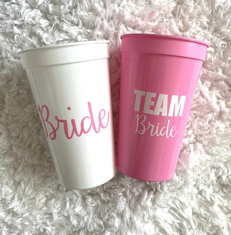 Puede incluir: Dos vasos de pl&aacute;stico, uno blanco y otro rosa. El vaso blanco dice "Bride" en letras rosas. El vaso rosa dice "Team Bride" en letras blancas.