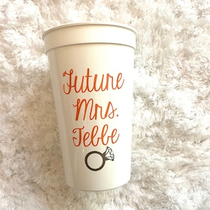May include: White plastic cup with the text "Future Mrs. [Name]" in orange lettering and a ring with a diamond graphic.