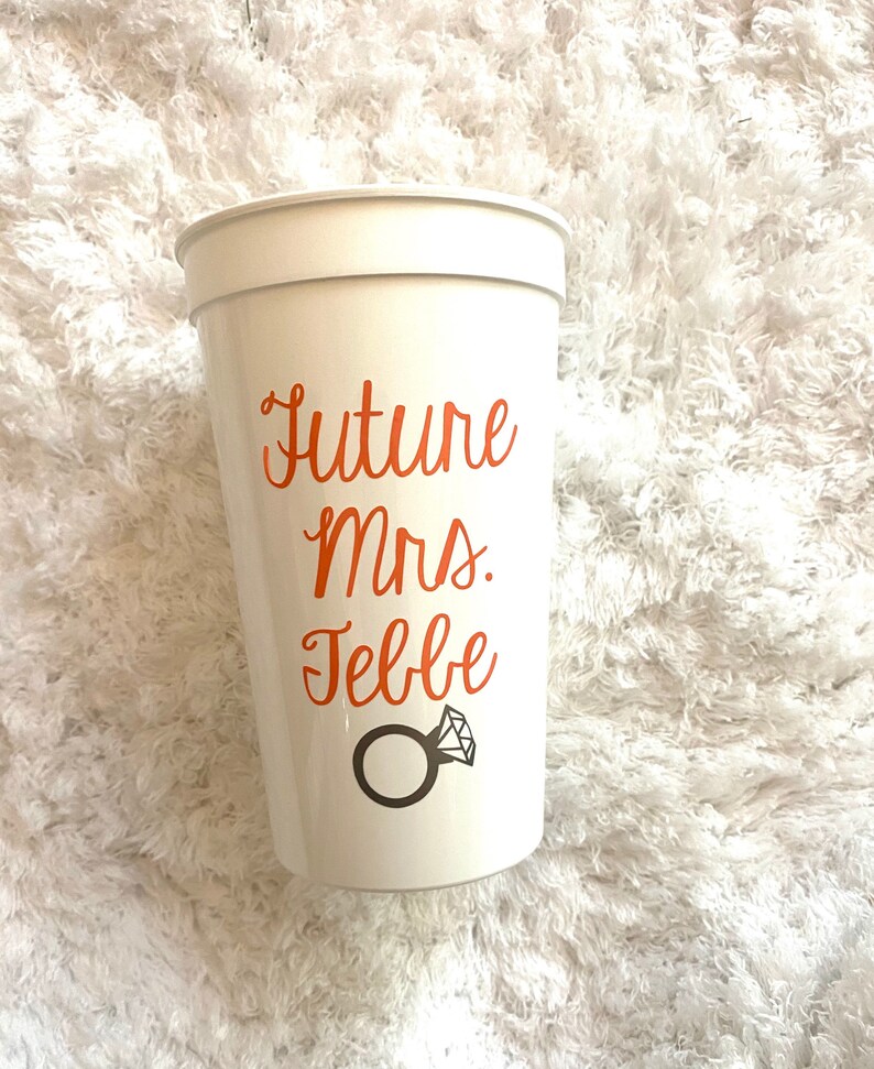 May include: White plastic cup with the text "Future Mrs. [Name]" in orange lettering and a ring with a diamond graphic.