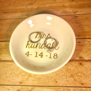 May include: White ceramic ring dish with gold lettering that reads "Mrs. Randall 4-14-18". Two silver rings are sitting on the dish.