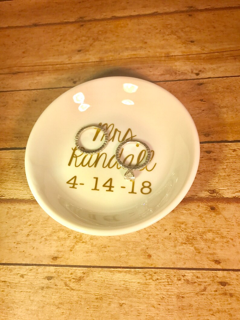 May include: White ceramic ring dish with gold lettering that reads "Mrs. Randall 4-14-18". Two silver rings are sitting on the dish.