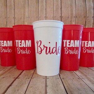 Puede incluir: Cinco vasos de pl&aacute;stico rojos con letras blancas que dicen "Team Bride" y un vaso de pl&aacute;stico blanco con letras blancas que dicen "Bride".