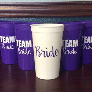 Puede incluir: Un conjunto de vasos de pl&aacute;stico morado con el texto "Team Bride" impreso en blanco en cada vaso. Un vaso es blanco con el texto "Bride" impreso en blanco.