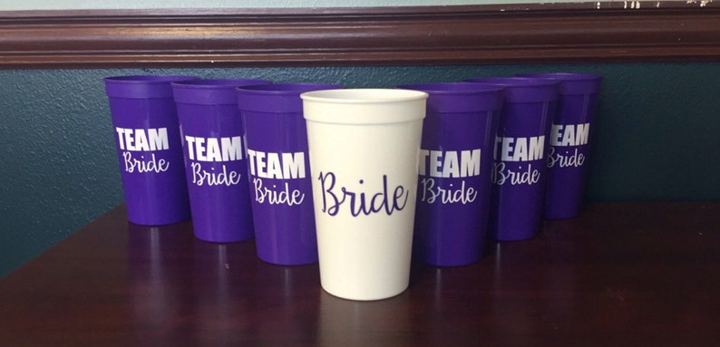 Puede incluir: Un conjunto de vasos de pl&aacute;stico morado con el texto "Team Bride" impreso en blanco en cada vaso. Un vaso es blanco con el texto "Bride" impreso en blanco.