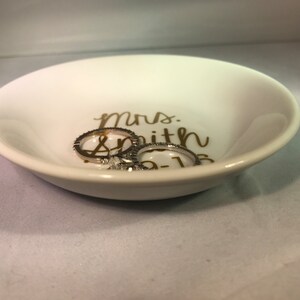 May include: White ceramic ring dish with gold lettering that reads "Mrs. Smith 05-16". Two silver rings are sitting in the dish.