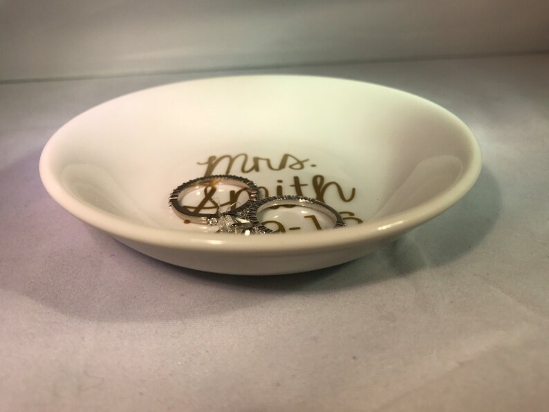 May include: White ceramic ring dish with gold lettering that reads "Mrs. Smith 05-16". Two silver rings are sitting in the dish.
