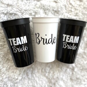 Puede incluir: Tres vasos de pl&aacute;stico con texto en blanco y negro. Un vaso es blanco con texto negro que dice "Bride". Los otros dos vasos son negros con texto blanco que dice "Team Bride".