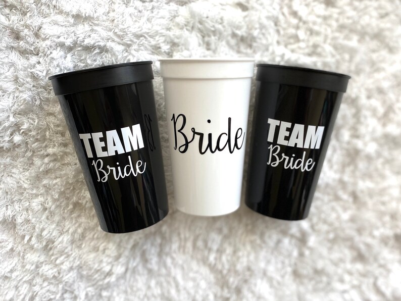 Puede incluir: Tres vasos de pl&aacute;stico con texto en blanco y negro. Un vaso es blanco con texto negro que dice "Bride". Los otros dos vasos son negros con texto blanco que dice "Team Bride".