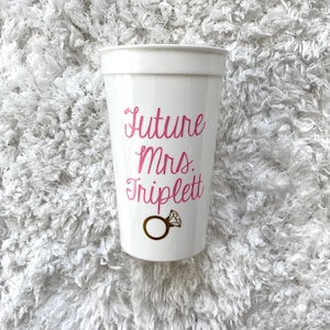 May include: White plastic cup with the text "Future Mrs. Triplett" in pink lettering and a gold ring with a diamond on it.