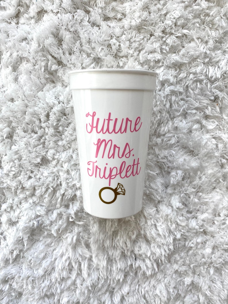 May include: White plastic cup with the text "Future Mrs. Triplett" in pink lettering and a gold ring with a diamond on it.