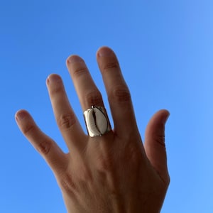 Peut inclure: Une bague en argent avec un motif de coquillage blanc. La bague est portée sur le majeur d'une main, la paume vers le haut.