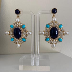 May include: A pair of ornate gold-toned earrings. Each earring features a large, dark blue oval center stone, surrounded by smaller turquoise stones, faux pearls, and clear crystals. A smaller dark blue stone tops each earring.