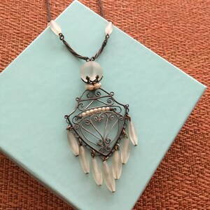 May include: An antique silver-toned necklace with a decorative pendant. The pendant features a filigree design with small pearls and teardrop-shaped glass beads. The necklace is displayed on a light blue box against a textured orange background.