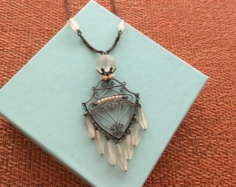May include: An antique silver-toned necklace with a decorative pendant. The pendant features a filigree design with small pearls and teardrop-shaped glass beads. The necklace is displayed on a light blue box against a textured orange background.