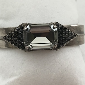 May include: A silver cuff bracelet with a large, clear, rectangular gemstone set in the center. The gemstone is surrounded by smaller black gemstones in a triangular shape on each side.