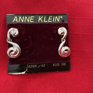 May include: A pair of silver earrings with a swirl design and a small clear stone accent. The earrings are displayed on a black card with the text "ANNE KLEIN" and "4286 / 42 $20.00".