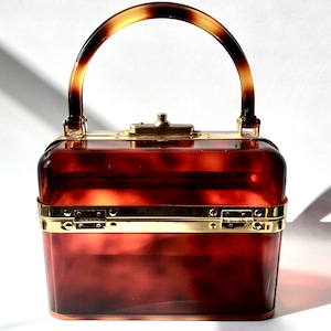 May include: A vintage-style, rectangular handbag with a tortoiseshell handle and a matching body. The bag features gold-colored metal accents around the edges and a clasp closure. The transparent body reveals a rich, reddish-brown hue.