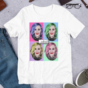 以下が含まれることがあります： カラフルなポップアートのデザインが施された白いTシャツ。4つの異なる髪色の女性が描かれています。Tシャツには「... bébé」という文字がプリントされています。
