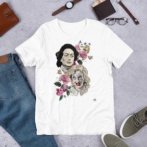 Pode incluir: Camiseta branca com um design floral com dois rostos de mulheres. As mulheres são representadas em um estilo vintage com flores rosa e brancas.