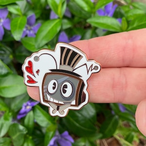Op de afbeelding: Een emaille pin in cartoonstijl met een retro televisiekarakter met een hoge hoed en antenne. De pin heeft een witte omtrek en een gouden rand. Het karakter heeft een lachend gezicht en rode hartaccenten. De pin is geplaatst tegen een achtergrond van groene bladeren en paarse bloemen.