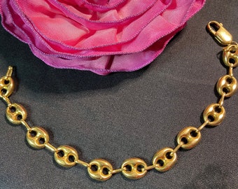 May include: A gold-toned chain bracelet with a mariner link design. The bracelet is on a black surface with a pink fabric flower.
