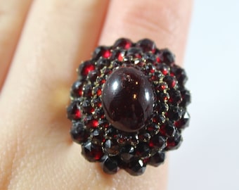 Op de afbeelding: Een rode granaten ring met een grote cabochon middelste steen omringd door kleinere granaten. De ring is gezet in goud.