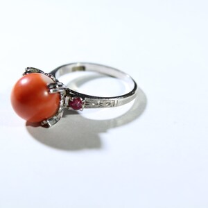 May include: A silver ring with a large, round, orange coral stone set in a prong setting. The ring is accented with two small red stones.