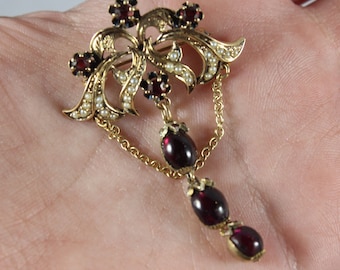 May include: A gold pendant brooch with a dangling chain and three red garnet stones. The top of the brooch features a floral design with red garnet stones and white pearls.