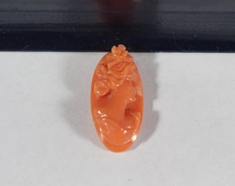 May include: An oval-shaped cameo carved from coral, featuring a detailed profile of a woman with intricate hair and clothing. The cameo is a vibrant orange-red color.