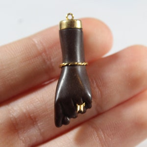 May include: A dark brown hand pendant with a gold band around the wrist and gold accents on the fingers. The pendant is made of wood and has a loop at the top for hanging.