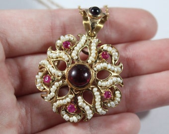 May include: A gold pendant necklace with a large, round, dark red gemstone surrounded by smaller, round, pink gemstones and white pearls. The pendant is in the shape of a flower with intricate details.