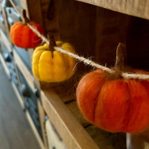 May include: A string of felt pumpkins in orange and yellow, hanging from a wooden shelf. The pumpkins are strung together with twine.