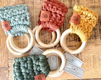 Puede incluir: Cuatro anillos de dentición de crochet en verde, naranja, amarillo y marrón con anillos de madera. Cada anillo tiene una etiqueta de cuero que dice "hecho a mano".