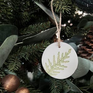 Op de afbeelding: Ronde witte ornament met een groen varenblad ontwerp. De ornament hangt aan een bruin touwtje aan een groene kerstboom.