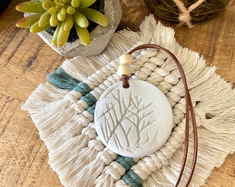 Peut inclure: Une ronde de céramique blanche avec un motif de branche d'arbre pend d'un cordon en cuir marron. L'ornement est sur une suspension murale en macramé blanc et turquoise avec des franges.