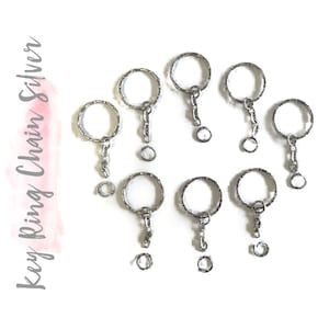 May include: A set of eight silver key rings with attached chains. The key rings are round and have a smooth finish. The chains are silver and have a simple design.