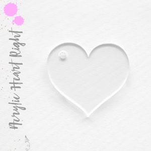 May include: A clear acrylic heart-shaped blank with a hole at the top. The heart is on a white textured background.