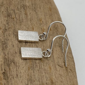 May include: A pair of silver dangle earrings with a rectangular shape. The earrings have a brushed finish and a simple design.