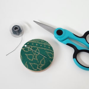 May include: A sewing kit with a green and tan patterned pin cushion, a spool of thread, and scissors with teal handles. A straight pin is inserted into the pin cushion. The scissors are open.