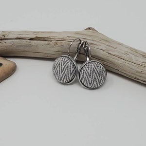 May include: A pair of round, silver-tone earrings with a black and white geometric pattern. The earrings have a lever-back closure and are displayed on a white surface with a piece of driftwood in the background.