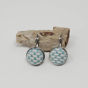May include: A pair of round, silver-toned earrings. Each earring features a light blue and white woven fabric pattern set within a circular frame. The earrings are displayed on a piece of light-colored wood against a white background.