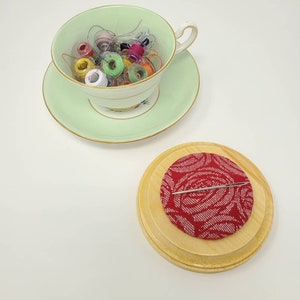 May include: A light green teacup and saucer filled with colorful sewing machine bobbins. A red pin cushion with a white floral pattern and a silver pin sits on a wooden base. The items are arranged on a white surface.