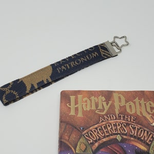 May include: A navy blue and gold wrist strap with the words "EXPECTO PATRONUM" and a stag design, next to a copy of "Harry Potter and the Sorcerer's Stone". The strap has a star-shaped metal clasp.