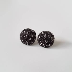 May include: Two round stud earrings. Each earring is a dark gray color with a woven white pattern. The earrings are set against a white background. The earrings are small and appear to be made of fabric.