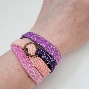 May include: A stack of four fabric bracelets in shades of pink, peach, purple, and navy blue, secured with a bronze-colored metal ring. The bracelets are wrapped around a wrist, showcasing their woven texture and color variations.