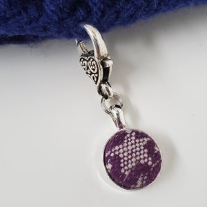 May include: A silver-toned charm with a heart-shaped clasp and a round pendant. The pendant features a purple and white patterned fabric design. The charm is attached to a section of dark blue knitted fabric.