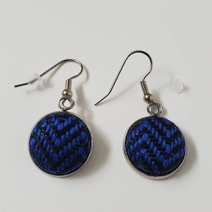 May include: A pair of round earrings with a blue and black woven fabric design. The earrings have a silver-colored metal frame and ear hooks. The earrings are shown on a white background.