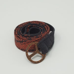 May include: A rolled-up belt with a copper-colored peace sign buckle. The belt is black with an orange and black woven pattern. The buckle is a stylized peace symbol.