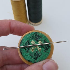 May include: A round wooden pincushion featuring a green fabric top with a black and beige geometric embroidered pattern. A long silver sewing needle is positioned horizontally across the center of the cushion. Two spools of thread are visible in the background.