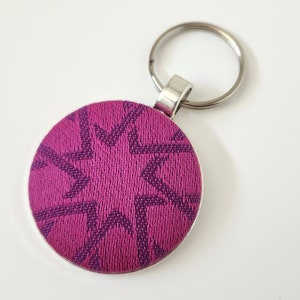 May include: A round keychain with a magenta fabric design featuring a dark purple star pattern. The keychain has a silver-colored metal border and a silver key ring.